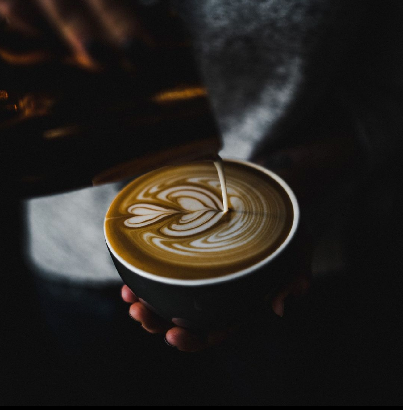 Close-up of a hand holding a mug as milk is poured to create intricate latte art. The dark, moody lighting emphasizes the swirling design.