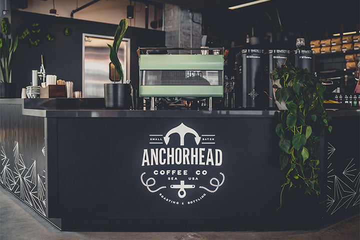 Sleek coffee shop counter with "Anchorhead Coffee Co" logo, featuring a mint green espresso machine, potted plants, and a modern, inviting atmosphere.
