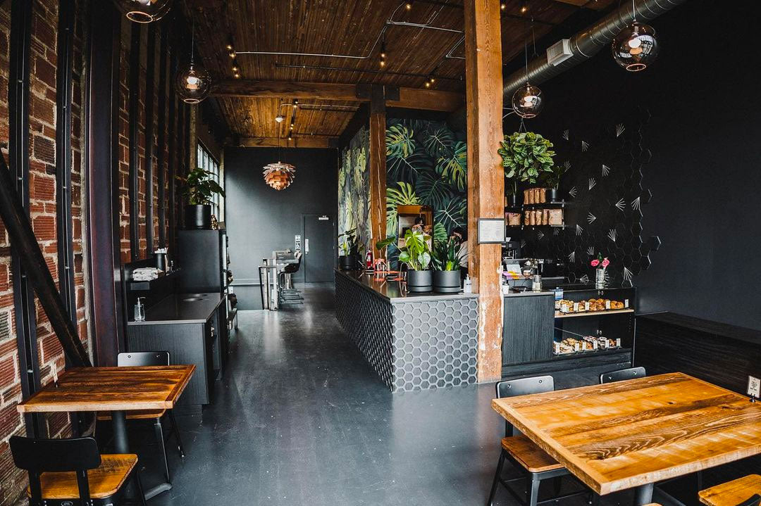 Cozy cafe interior with wooden tables and chairs, brick walls, potted plants, and black hexagonal tiles on the counter. Warm, inviting atmosphere.