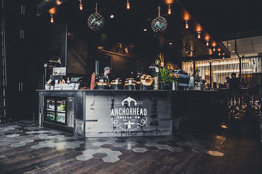 Trendy coffee shop with dim lighting and industrial decor. The counter displays pastries and plants, with a barista working behind. Modern, cozy ambiance.