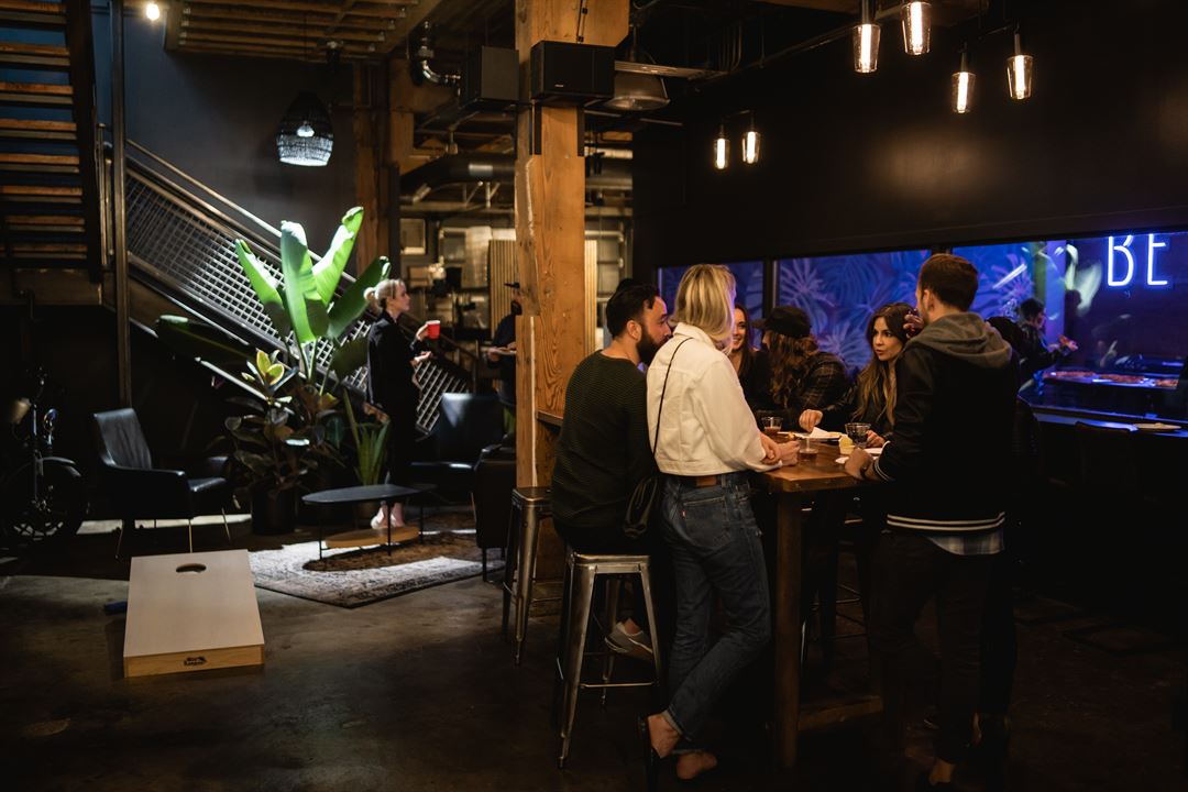 A cozy bar setting with a group of people chatting around a high table. Dimmly lit, warm ambiance. Background shows plants, seating, and a neon sign.