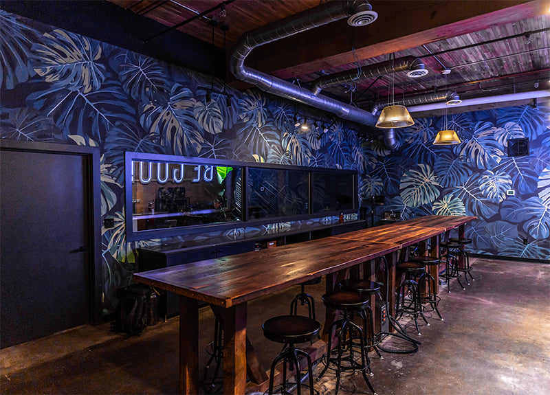 Modern bar with a long wooden table and stools. Blue leaf-patterned walls and exposed pipes create an industrial-chic vibe. Dim, warm lighting.